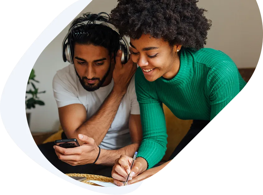 Happy woman writing notes while her husband applies for a loan with guaranteed approval on his phone